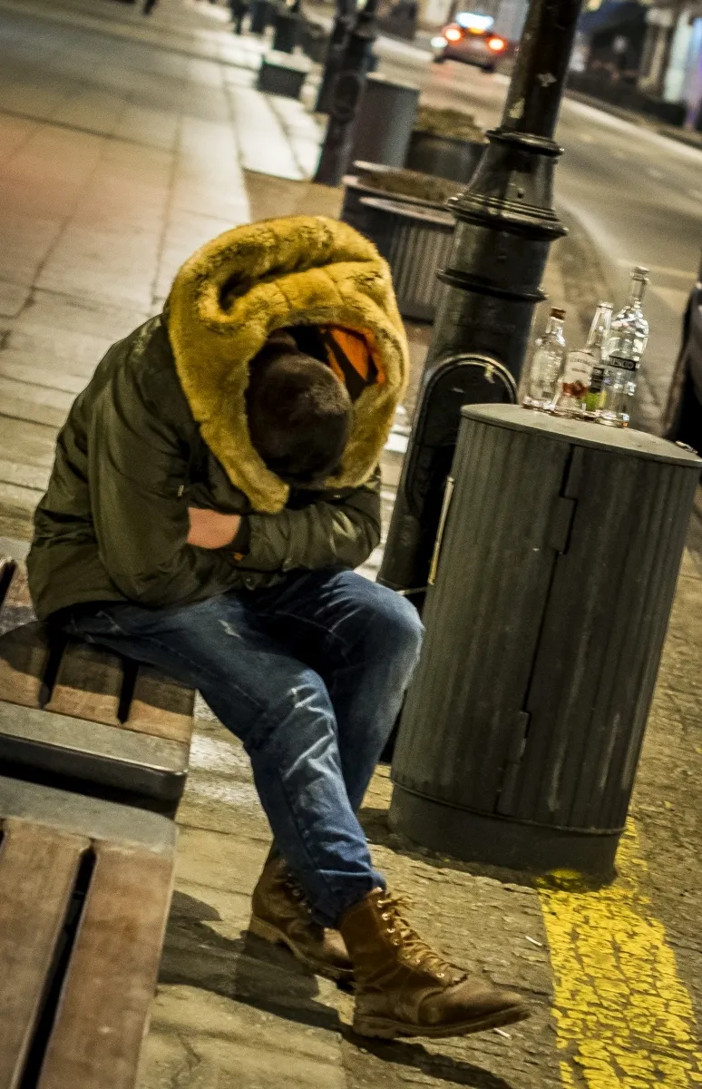 Seated man curled inside a fur-lined hood on a nighttime sidewalk beside empty bottles